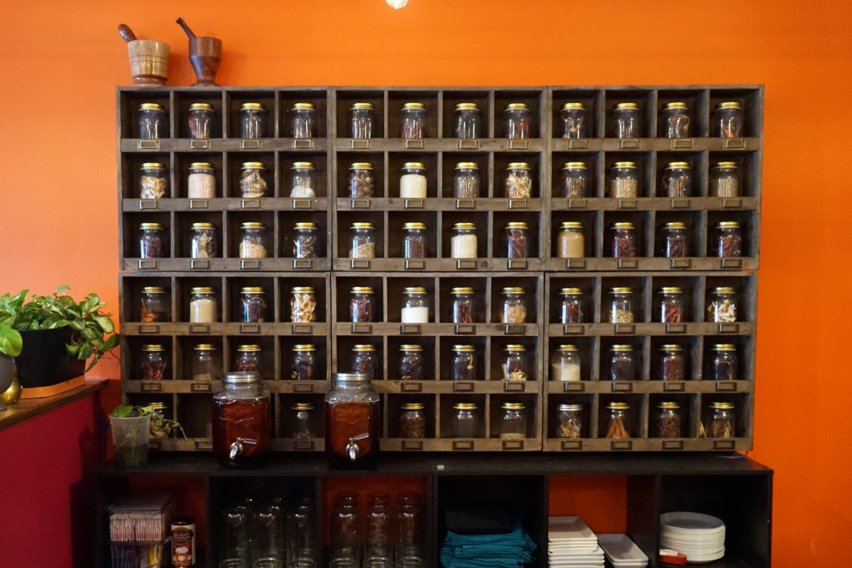 A photo of a big wooden spice rack full of jars that is hanging from an orange wall in a restaurant.