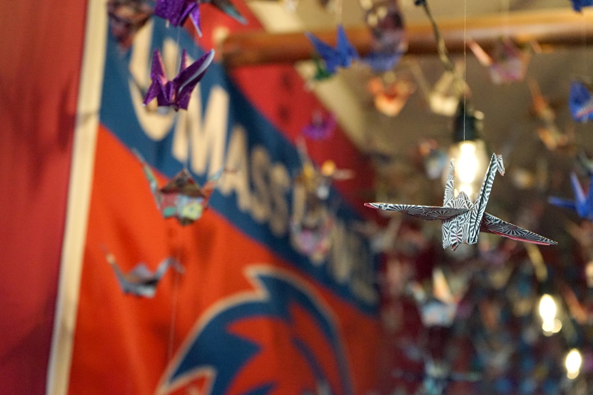 Dozens of paper cranes hang from a ceiling. There is a UMass Lowell flag hanging on the wall in the background.