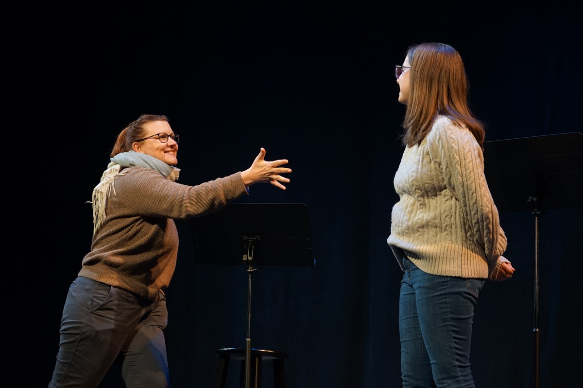 A woman in glasses with a scarf around her neck gestures with her right hand toward a woman on a stage.